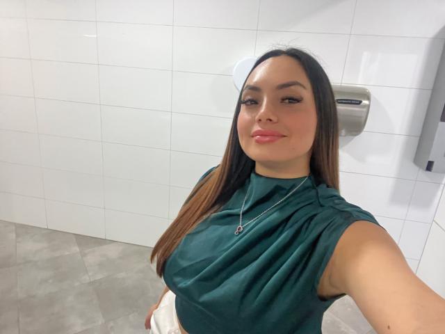 GalaMalkova, avec de longs cheveux bruns, un haut vert sans manches, du rouge à lèvres rose et un collier, se prend en photo dans une pièce carrelée, montrant légèrement sa poitrine, et souriant avec assurance..