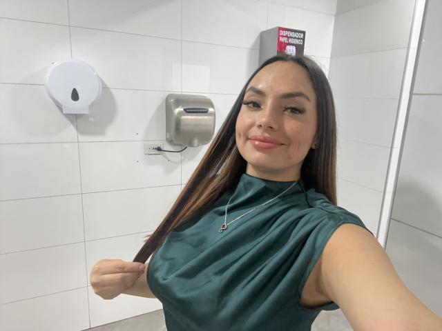 GalaMalkova with long brown hair, wearing a green sleeveless top, lifting her right breast with her left hand, smiling slightly, in a white-tiled bathroom.