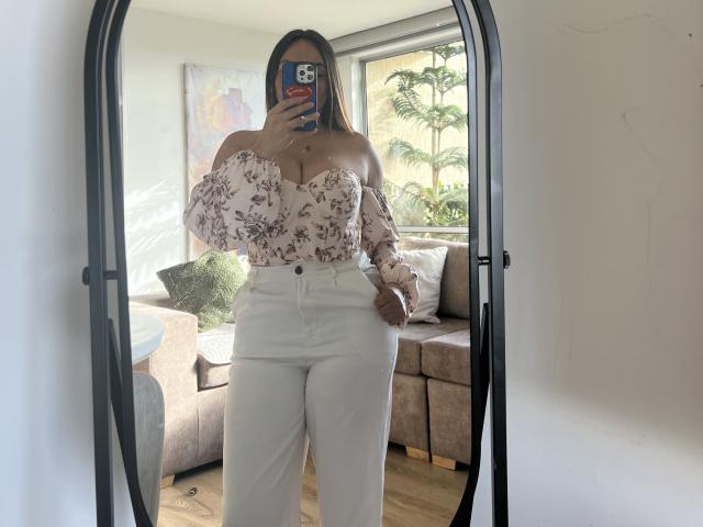 GalaMalkova, avec de grandes seins, porte un haut à fleurs sans manches et un pantalon blanc, et prend un selfie devant un miroir, tenant un téléphone avec une coque colorée, dans une pièce baignée de soleil..