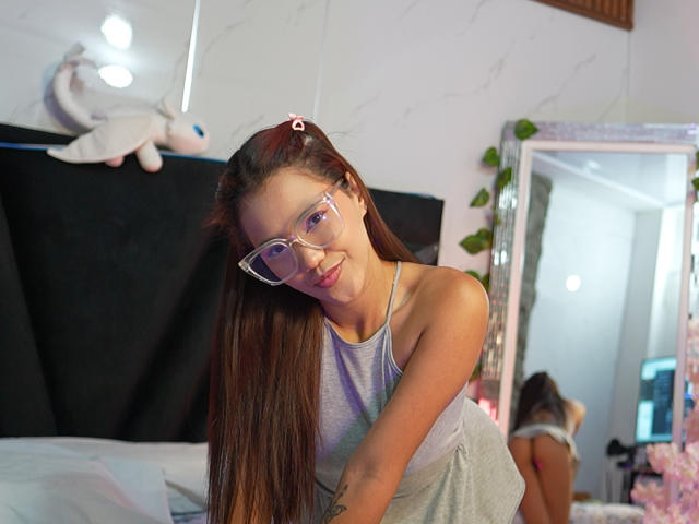SkinnySweetMilff with long brown hair, wearing clear glasses and a grey tank top, leaning forward, smiling, showing her small tits, in a brightly lit room with a mirror.