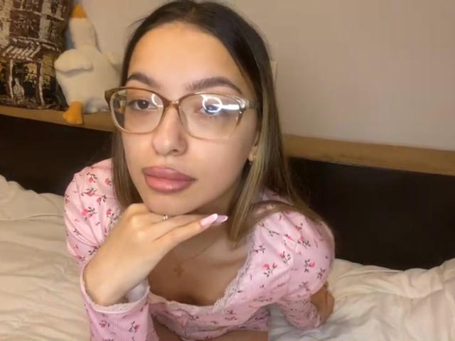 RaneeDerienzo with glasses, pink floral top, and pink nails, resting chin on hand, showing tits, on bed, penguin stuffed toy in background.