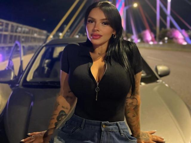 CarlaSuarez69 con grandi seni, top nero a coste, zip, pantaloncini di jeans, tatuaggi, lunghi capelli neri, rossetto rosa, in piedi accanto a un'auto di notte, che guarda verso la fotocamera..