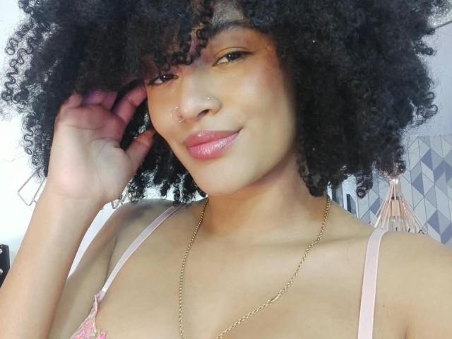SnackTime a les cheveux noirs bouclés, une peau brun clair, un anneau au nez et du rouge à lèvres rose. Elle porte un soutien-gorge rose avec un collier en chaîne dorée, et sourit doucement, avec une main près de son visage..