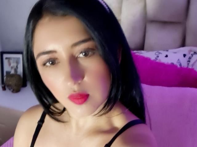 NicollJeans has black hair, red lipstick, and is wearing a black spaghetti strap top, with her face centered in the image, looking slightly to the left.