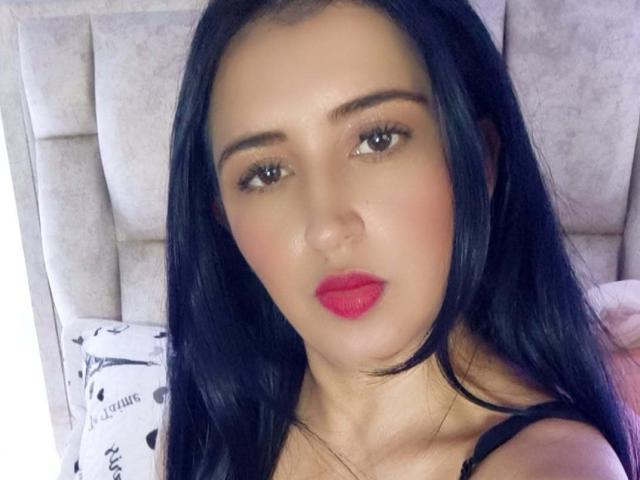 NicollJeans has black hair, brown eyes, red lipstick, and a cute face with slightly flushed cheeks. She's wearing a black strap top, looking at the camera.
