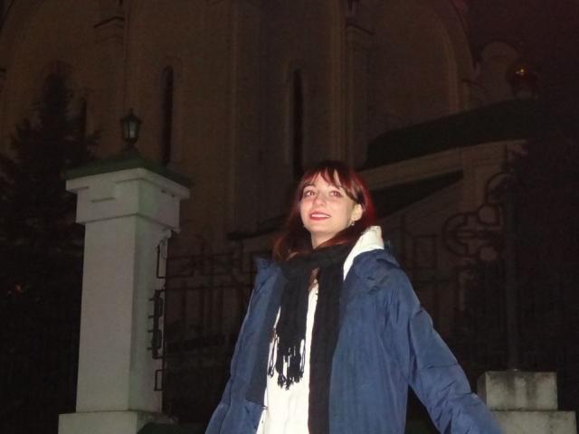 LeomaHaq with red hair, blue coat, black scarf, white dress, standing outside at night, looking up with a slight smile, dark background, and a white pillar to her left.