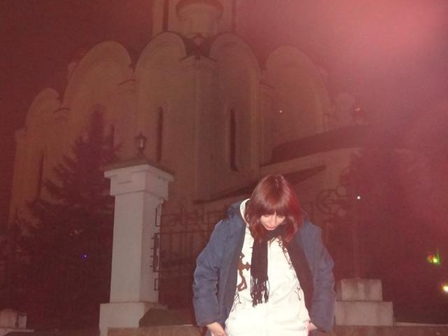 LeomaHaq with brown hair, wearing a blue jacket and white dress, black scarf, standing in front of a stone building at night, looking down, red light flare in the background.