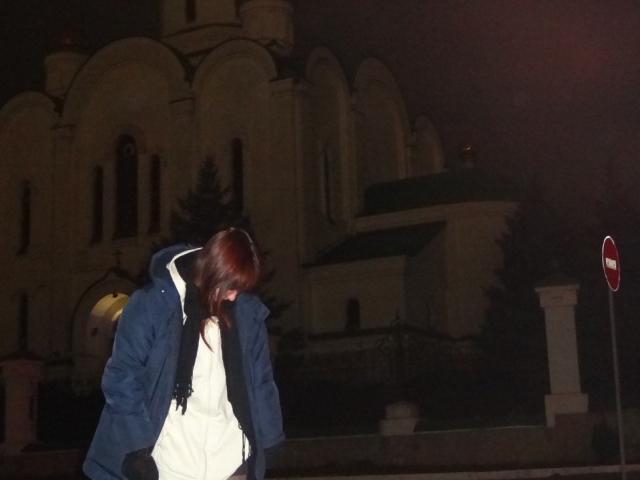 LeomaHaq with long red hair, wearing a blue jacket and white hoodie, bending forward, hands in pockets, at night, in front of a church with a stop sign.