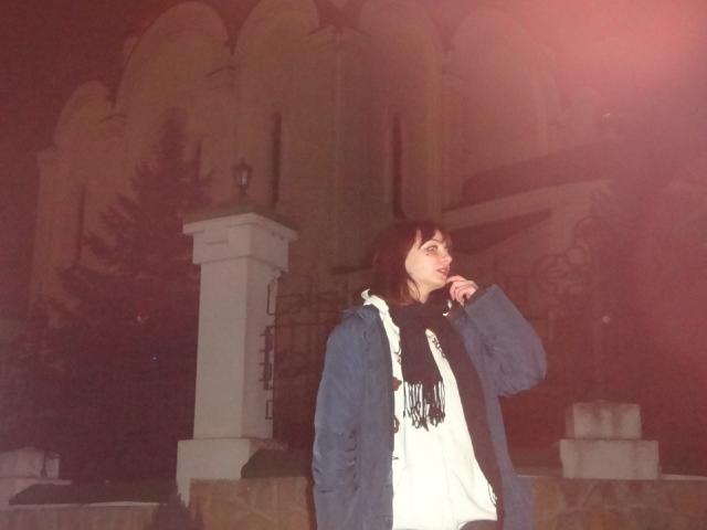 LeomaHaq stands in the night, wearing a blue jacket, white shirt, and black scarf, with her hand near her mouth, looking to the side. Bright light in the background.