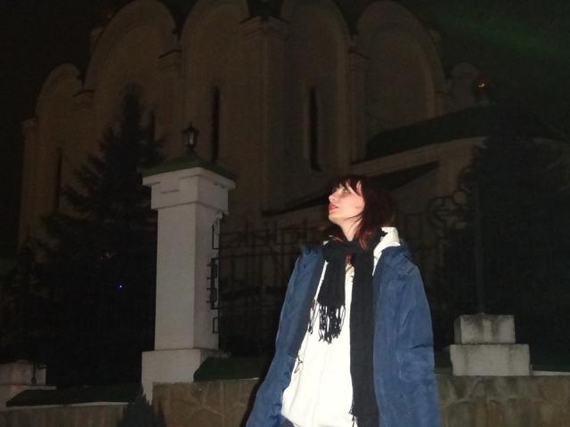 LeomaHaq stands at night, wearing a blue jacket, white dress, and black scarf, looking up with a slightly open mouth, dark brown hair, and fair skin.