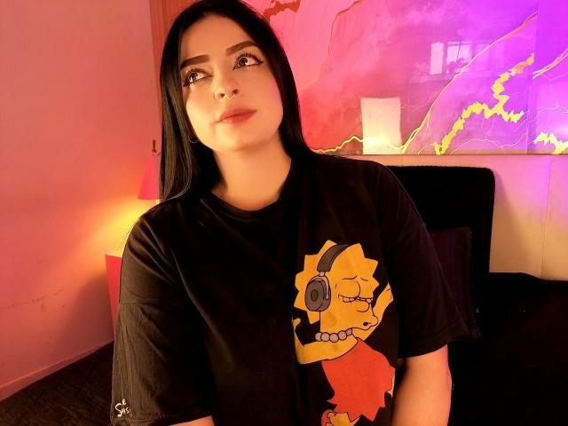 LilithAmoth with long black hair, wearing a black t-shirt with a yellow cartoon character, looking up with a slight smile, in a warmly lit room.