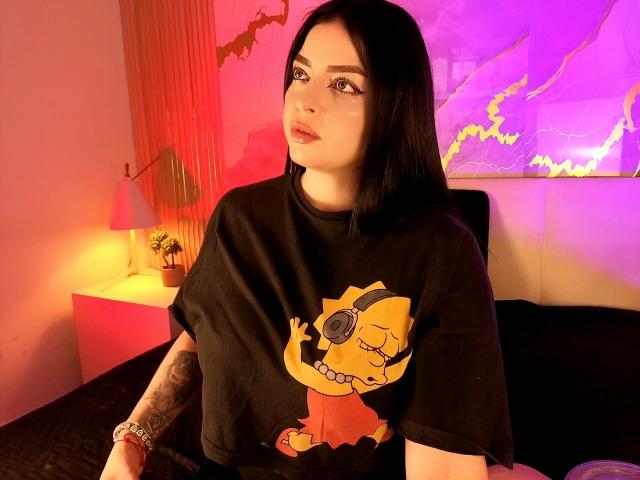 LilithAmoth with black hair, wearing a black The Simpsons t-shirt with Bart wearing headphones, sitting on a bed, tattooed arm, pink and purple lighting, looking at camera.