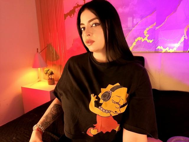 LilithAmoth with long black hair, pale skin, tattoos on right arm, wearing a black shirt with a yellow cartoon character, sitting in a room with pink and purple lighting.
