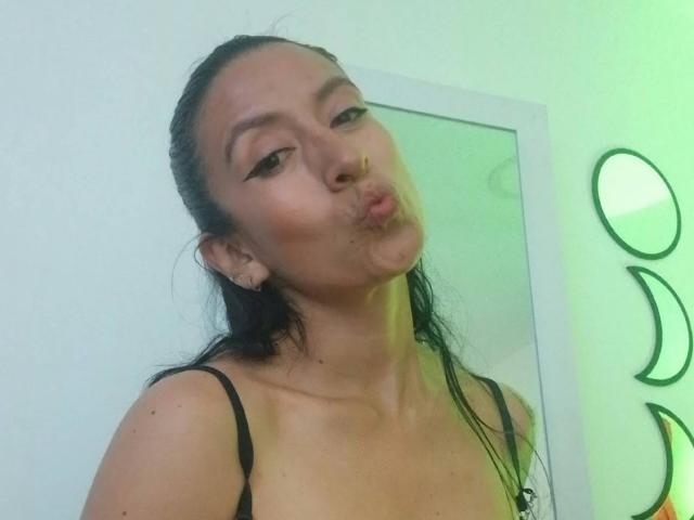 ZarayFeet with wet black hair, puffy lips making a kissy face, wearing a black spaghetti strap top, showing small tits, standing in a green-lit room.