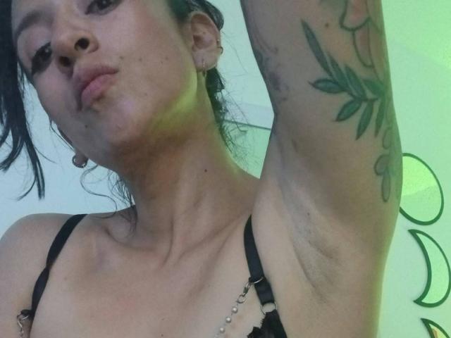 ZarayFeet con labbra piene, reggiseno nero, braccio sollevato che mostra un tatuaggio a forma di foglia, piercing alle orecchie e leggera peluria sotto le ascelle, luce verde sullo sfondo. Donna asiatica, carnagione media..