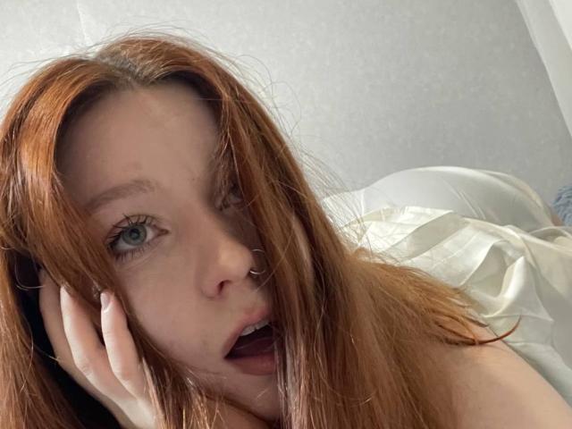 LilliamChevalier has long red hair, fair skin, and blue eyes, with slightly parted lips and her hand touching her face in a sleepy, sensual manner.