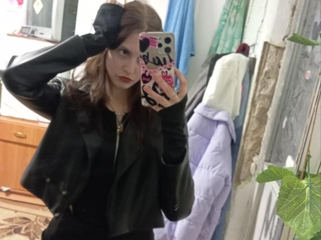 EddaPack mit braunen Haaren, einer schwarzen Lederjacke und rotem Lippenstift, die ein Selfie vor einem Spiegel macht, wobei sie mit einer Hand ihr Haar berührt. Sie hält ein Telefon mit einer rosa Aufkleberhülle..