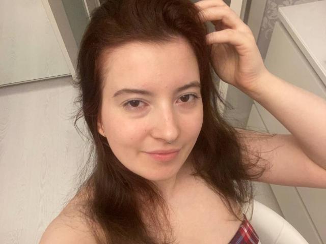JohnnaChelf, con lunghi capelli castani, pelle chiara e occhi marroni, sorride leggermente, tenendosi i capelli con una mano, indossa una camicia a quadri rossa, in una stanza bianca..