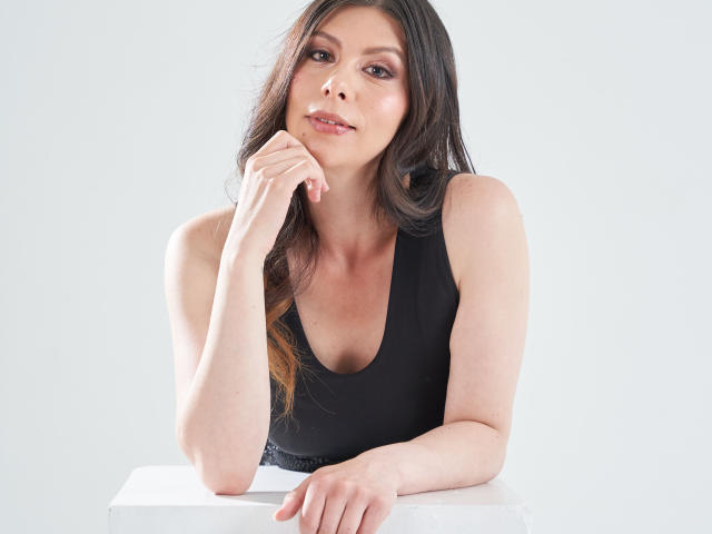KarlaMazuera with long brown hair, wearing a black tank top, resting her chin on her hand, looking slightly to the side with a subtle smile, white table in front.