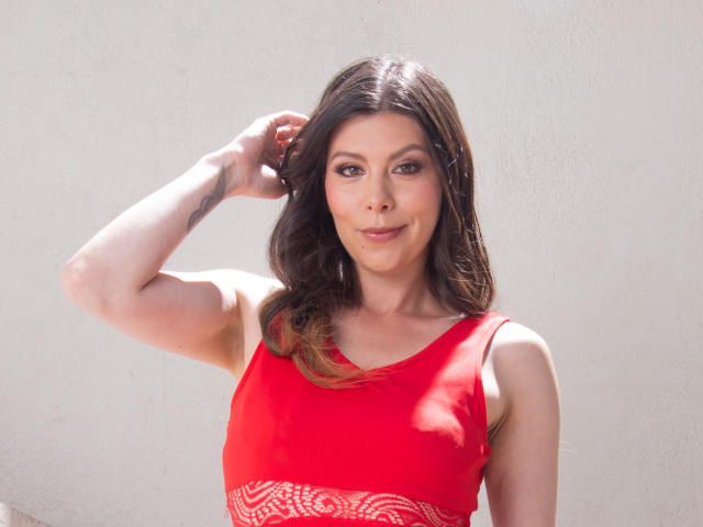 KarlaMazuera with brown hair, red dress, and small tattoo on her arm, raising her right hand, smiling, showing her tits, and looking at the camera.
