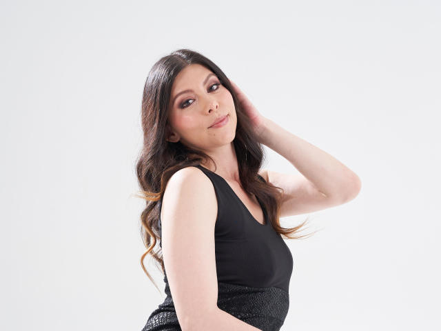 KarlaMazuera has long brown hair, black dress, and is looking at the camera with a slight smile, hand in hair, showing off her tits. She's cute and sexy.