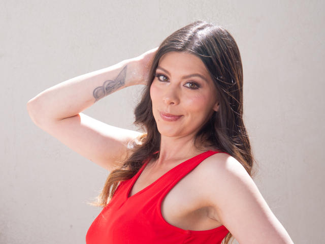 KarlaMazuera with long brown hair, red dress, smiling, arm up showing tattoo, light skin, brown eyes, slight makeup, sunlight on face, white wall background, soft expression.