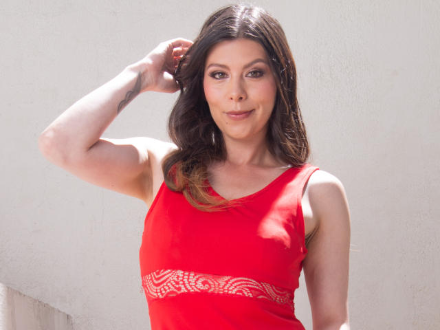 KarlaMazuera with brown hair, red sleeveless top, and white pattern, raising arm, smiling, showing small tits, tattoo on forearm, light skin, standing against plain wall.