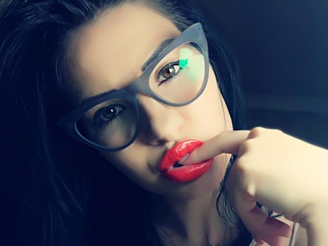 Zaccada with dark hair, wearing glasses, biting her red lipstick-covered finger. Her green eyes are slightly squinted. She's looking seductive with her hand near her mouth.