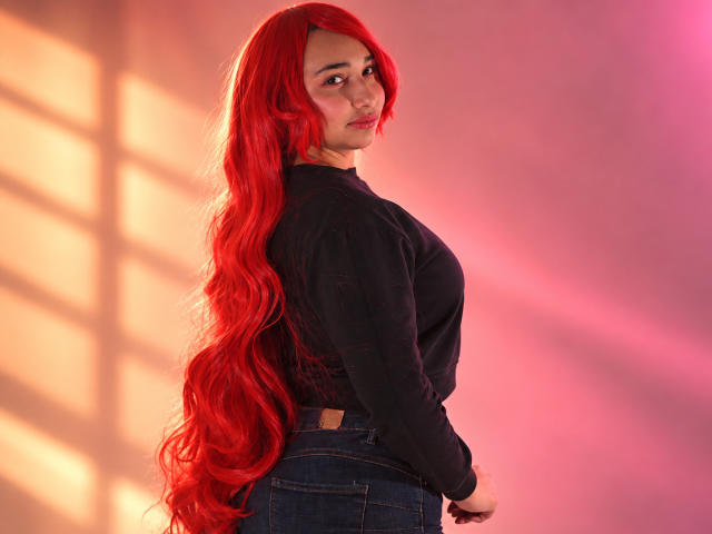 EleanorLamberth with long red hair, black shirt, and blue jeans, looking over her shoulder, showing her ass, in a pink and orange lit room.