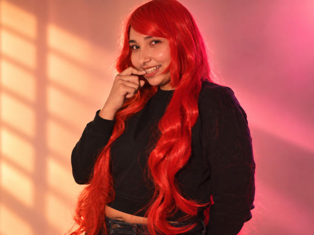 Eleanor Lamberth mit einer langen roten Perücke, lächelnd, die Nase berührend, trägt ein schwarzes Oberteil mit langen Ärmeln, steht, rosa und orangefarbenes Licht, Schatten an der Wand, süß, sexy..