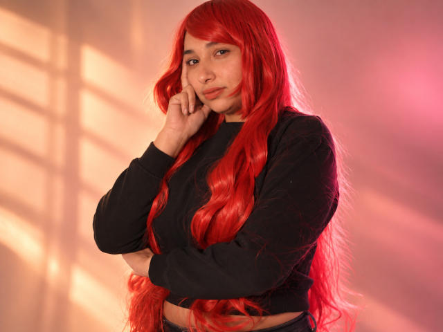 Eleanor Lamberth mit roter Perücke, langem, gewellten Haar, schwarzem Crop-Top und schwarzen Ärmeln, die ihre Wange berührt, stehend mit verschränkten Armen, rosa und orangefarbenes Licht..