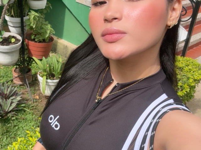 MiiaPalmer with shiny pink blush, black sleeveless top, and gold necklace, showing her tits in a garden, black hair, pink lips, and gold earrings.