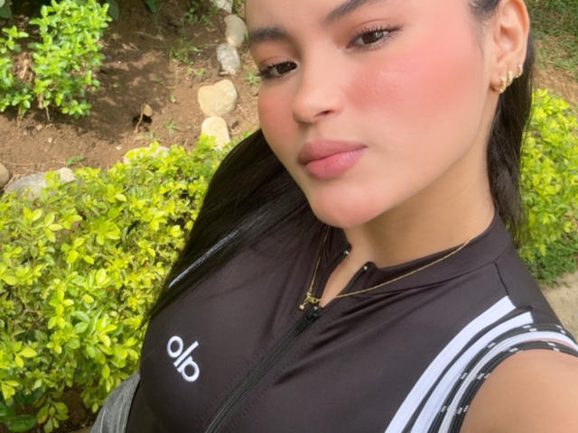 MiiaPalmer has pink blushed cheeks, full lips, and long black hair. She's wearing a black athletic top with white stripes, showing her small tits. She's looking at the camera.
