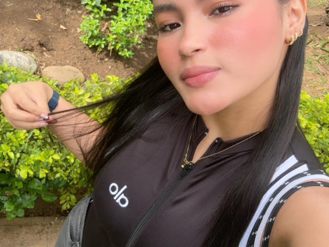 MiiaPalmer with black hair, pink cheeks, and a slight smile, holding her hair, wearing a black Adidas sports top, showing her small tits, standing outdoors.