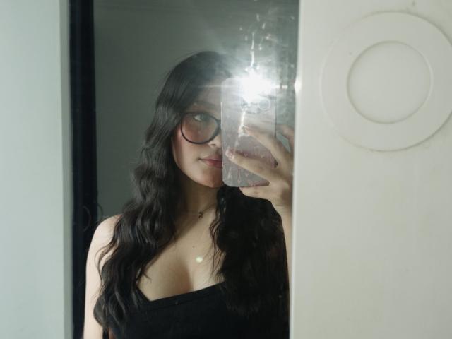 MiiaPalmer with long wavy black hair, glasses, and a low-cut black top showing her tits, taking a mirror selfie with a flash on her phone.