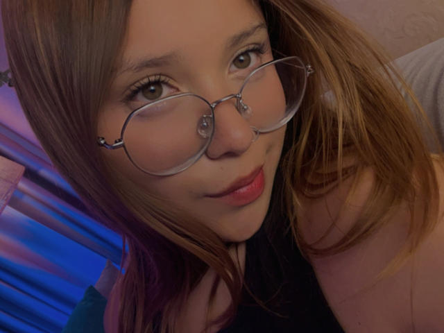 HeraJuno, usando óculos, com cabelo castanho e lábios vermelhos, sorrindo para a câmera. Seus seios estão ligeiramente visíveis na parte inferior da imagem. Ela tem um rosto bonito e atraente..