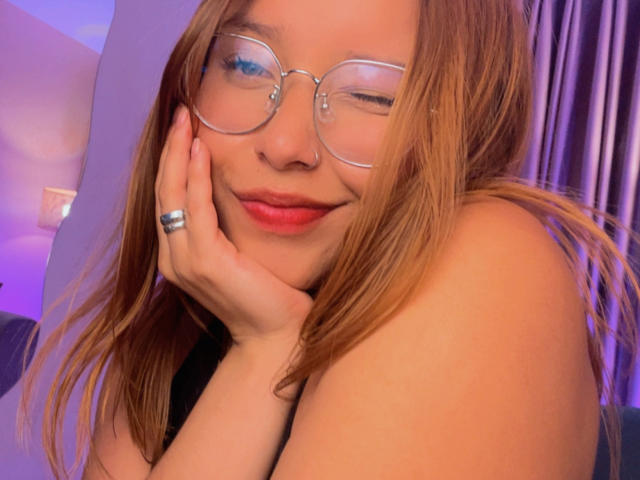 HeraJuno avec des lunettes, clignant de l'œil, avec des lèvres rouges, touchant sa joue, montrant ses seins, et portant une bague en argent, dans un autoportrait avec un éclairage violet..