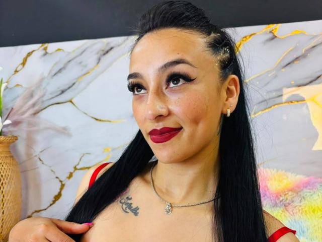 Scarlett Pinzón, con cabello negro largo, lápiz labial rojo y delineador de ojos oscuro, vestida con una blusa roja, mostrando senos pequeños, un tatuaje en el pecho, un collar y aretes, sonriendo ligeramente..