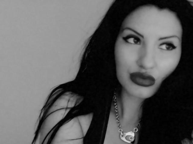 Black-and-white photo of Zaccada with long dark hair, thick eyebrows, dark lipstick, looking to the side, wearing a chain necklace, showing bare shoulders. Zaccada's tits are not visible.