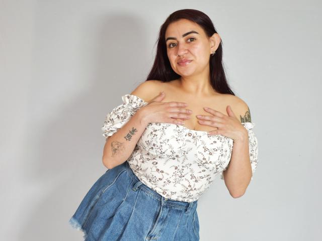 MarianaRusell with dark hair, off-shoulder white floral top, blue high-waisted jeans, hands on her tits, tattoo on right arm, smiling, light gray background, earrings.