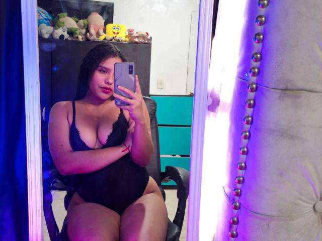 AriaVega with big tits, wearing a black lace bodysuit, sitting in a chair, taking a mirror selfie with her phone, in a room with purple LED lights.