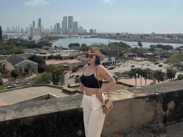 IsabellaJames, con la pelle abbronzata, reggiseno sportivo nero che lascia intravedere piccoli seni, pantaloni bianchi, cappello da cowboy, occhiali da sole, tatuaggio sul braccio sinistro, che tiene una borsa dorata, in piedi su un muro di pietra, con lo skyline della città sullo sfondo..