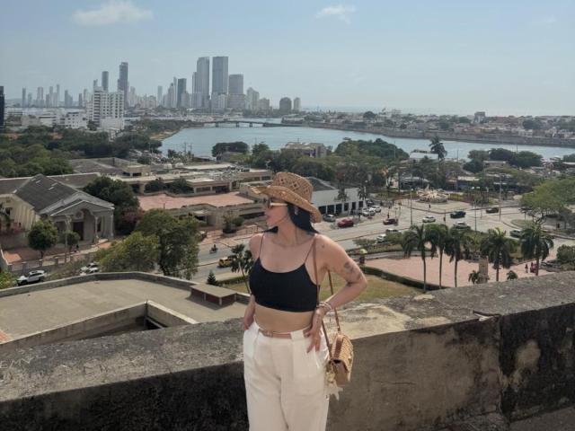 IsabellaJames stands with hands on hips, wearing a black crop top, white high-waisted pants, and a straw hat, holding a tan purse, with a city skyline in the background.