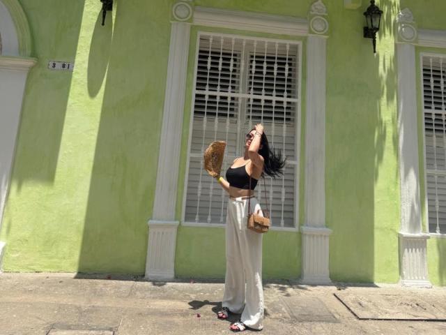 Isabella James, vêtue d'un soutien-gorge noir et d'un pantalon blanc, se tient sur une rue ensoleillée, tenant un éventail en osier, les yeux fermés, un sac en toile marron à ses côtés, de longs cheveux noirs, une peau bronzée et des sandales rouges..