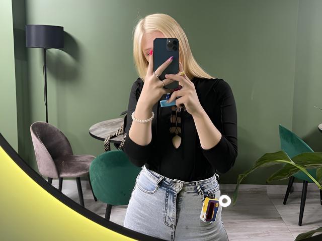 KerstinBrose with blonde hair, black top, and high-waisted jeans, taking a mirror selfie, showing pink lipstick, blue nails, and a ring, with a phone and a bracelet.