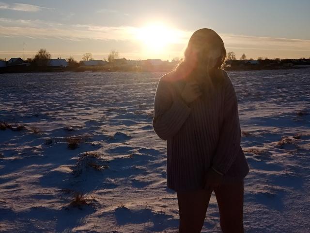 KerstinBrose stands in a snowy field, sun setting, wearing a loose, purple, rib-knit sweater, covering her small tits with one hand, shadowing her face, sunlight in her hair.