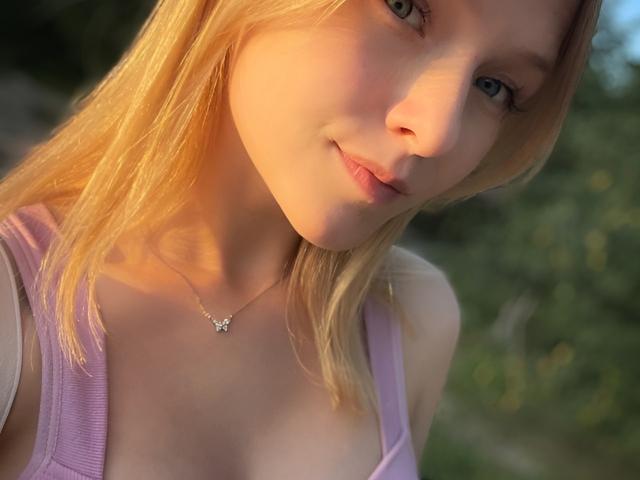 KerstinBrose has blonde hair, light skin, and small tits, wearing a purple top, a butterfly necklace, smiling at the camera in a sunny outdoor setting.