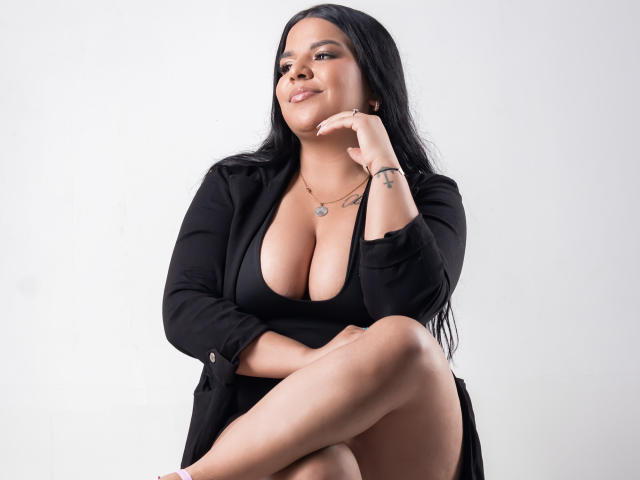 GabbyCastello with large boobs, sitting, wearing black shirt, deep cleavage, long black hair, confident smile, hand on chin, crossed legs, tattoo on wrist, white background.