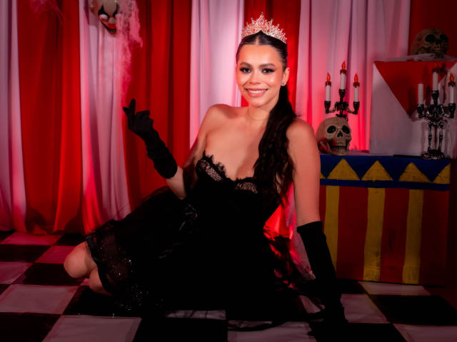 Alana Blake mit dunklem Haar, zu einem Zopf geflochten, trägt ein schwarzes Spitzenkleid, schwarze Handschuhe und eine silberne Krone. Sie kniet auf einem Schachbrettmuster-Boden und lächelt in die Kamera..