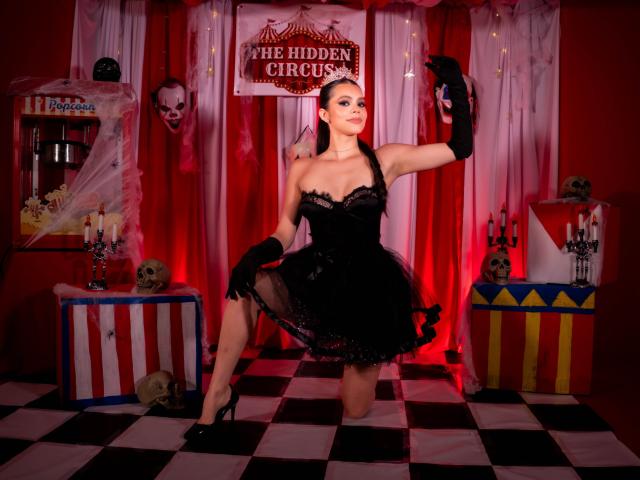 Alana Blake trägt ein schwarzes Spitzenkleid, Handschuhe und High Heels, kniet und zeigt ihr Gesäß. Sie befindet sich in einer Zirkus-ähnlichen Umgebung mit Schädeln, Masken und roter Beleuchtung. Sie hält eine blaue Karte..
